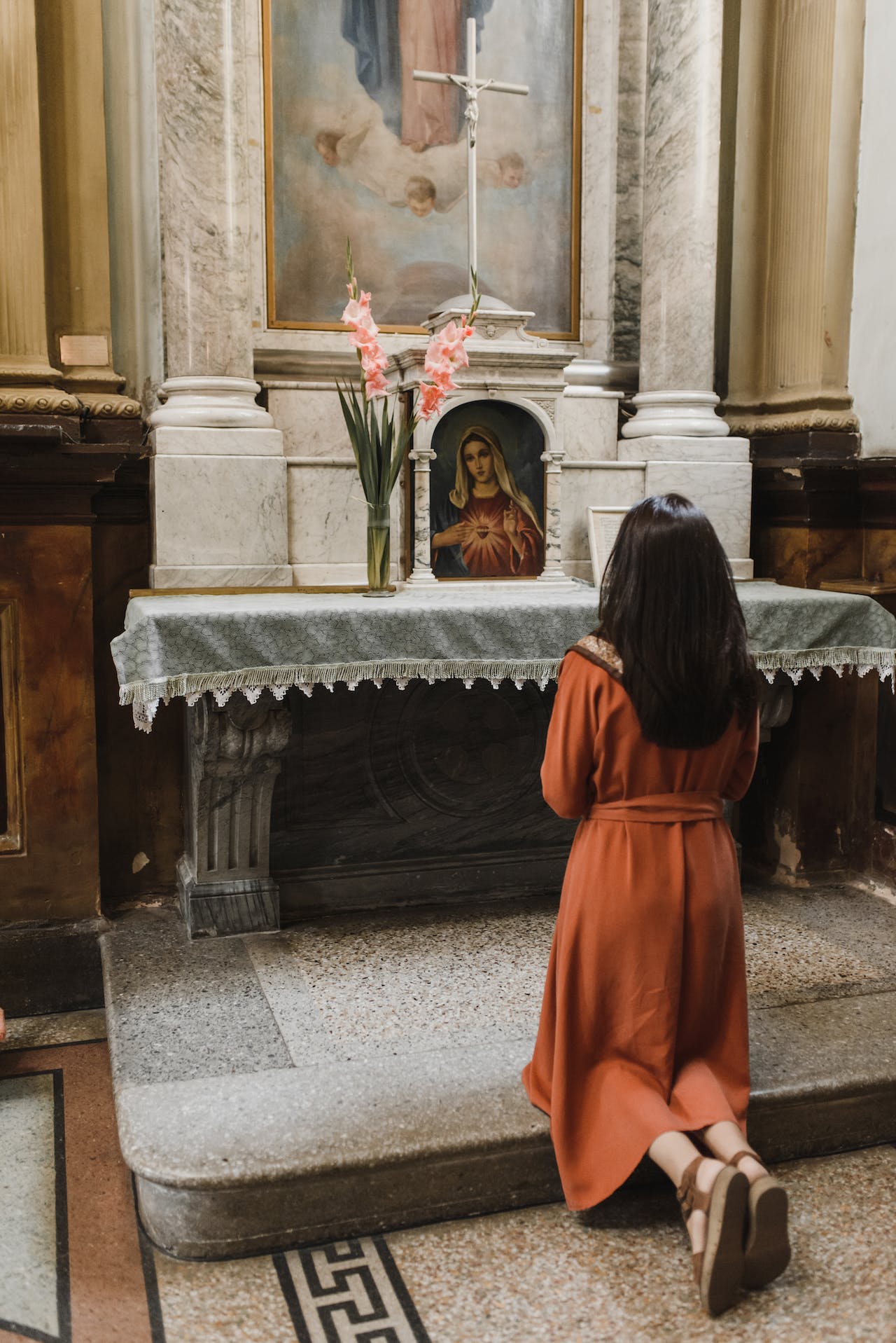 Mulher ajoelhada diante de um crucifixo e da imagem de Nossa Senhora